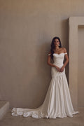 Woman in a white off-shoulder wedding dress standing against a beige wall.